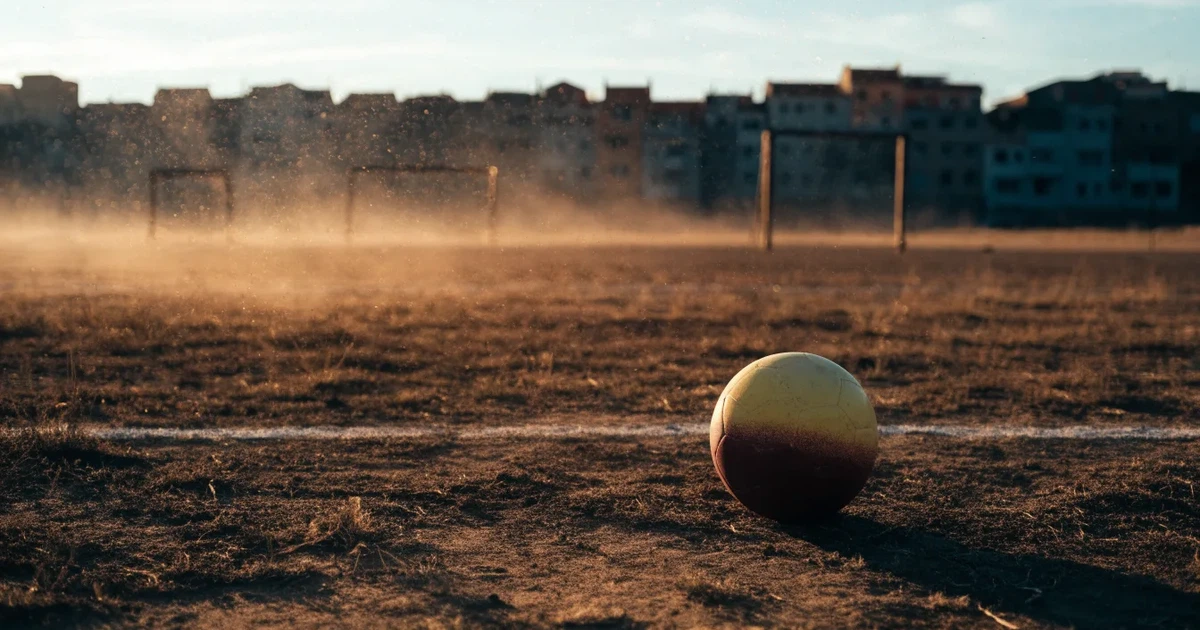 Ein sandiger Fußballplatz am Rand einer Stadt in warmem Abendlicht, Staub in der Luft und zwei Tore in der Ferne.