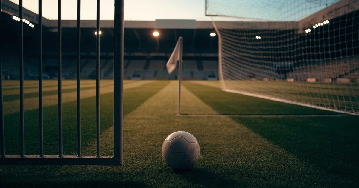 Blick durch ein Stadiontor auf einen grünen Rasen im Abendlicht, ein Ball ruht an der Eckfahne, die Tribünen sind leer.