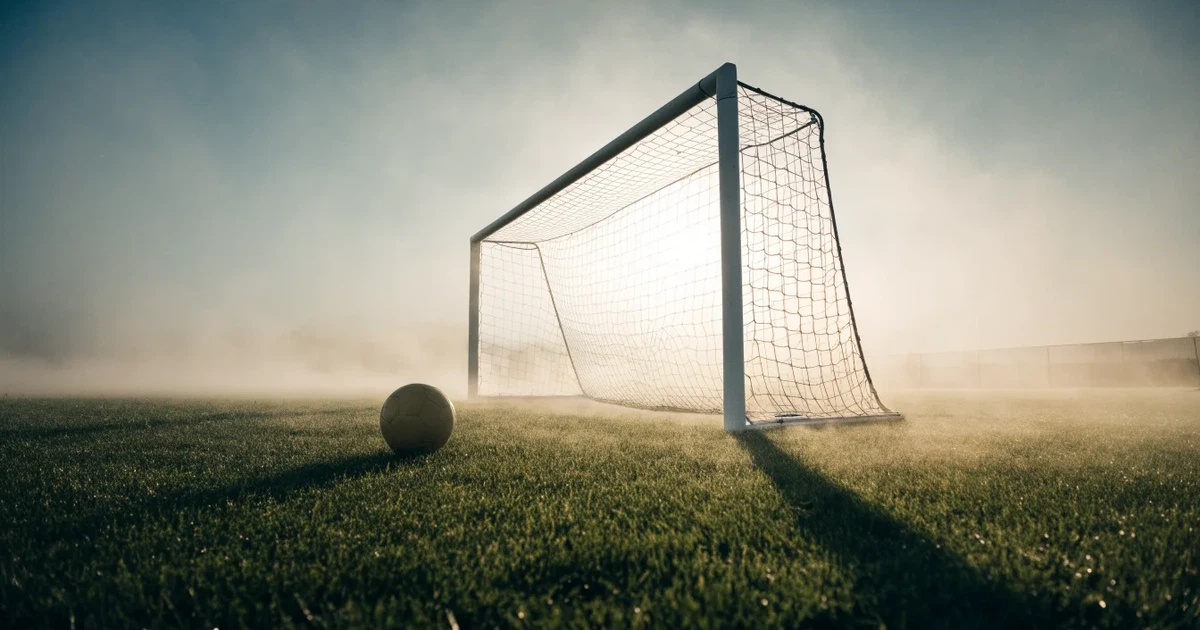 Ein Fußballtor mit gespanntem Netz steht auf einem leeren Rasenplatz, davor ein einzelner Ball, Morgendunst liegt über dem Feld.