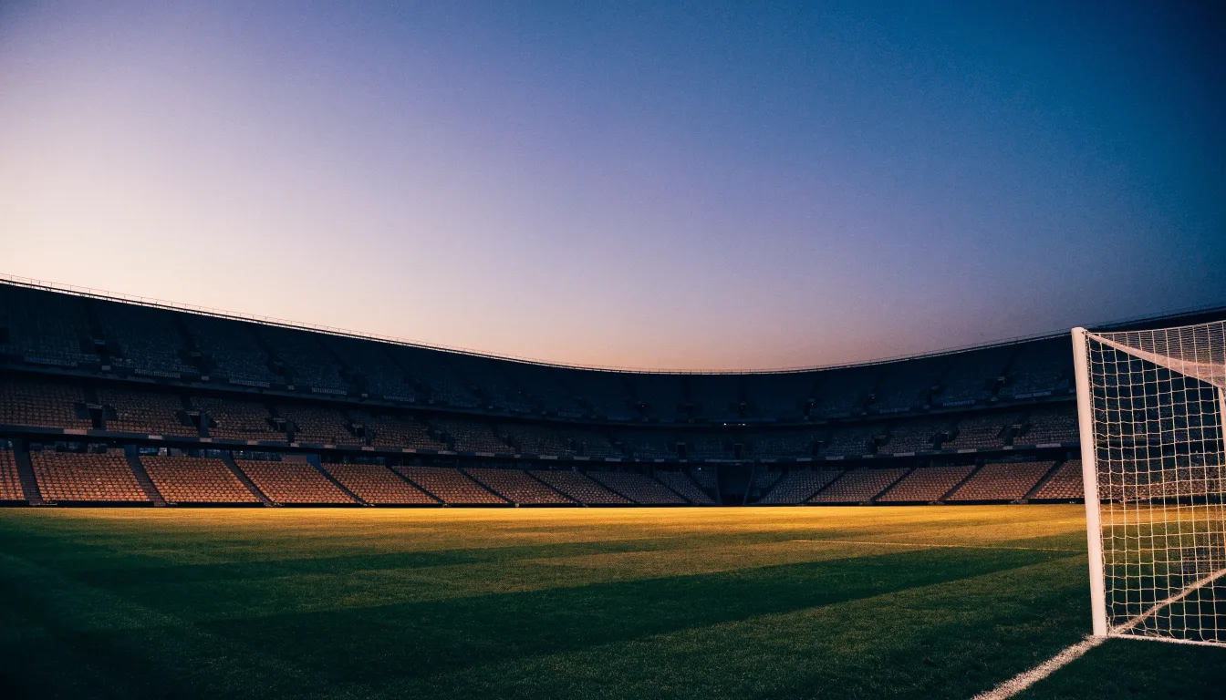 Eine leere Stadiontribüne unter Flutlicht aus der Ferne betrachtet, die Rasenfläche leuchtet grün in der Dämmerung.