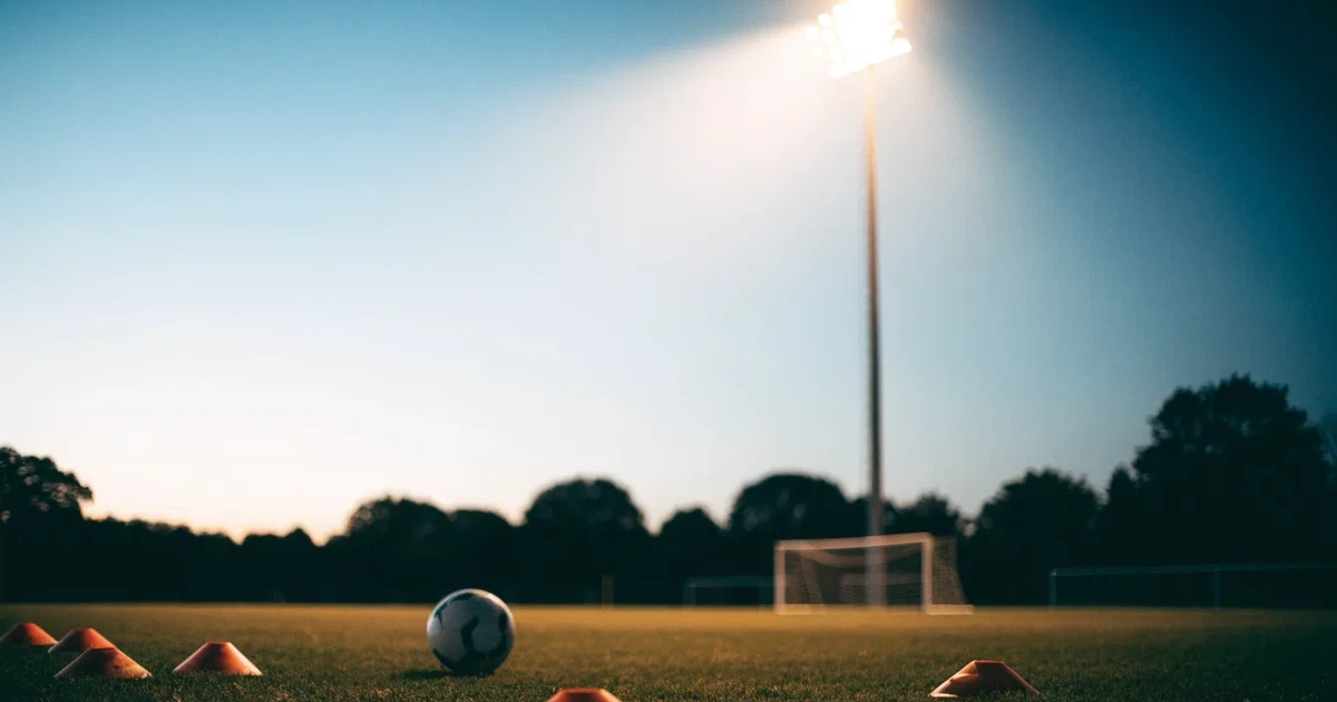 Eine Flutlichtanlage ragt über einen Trainingsplatz, auf dem Rasen liegen verstreut Hütchen und ein Ball, am Himmel die Abenddämmerung.