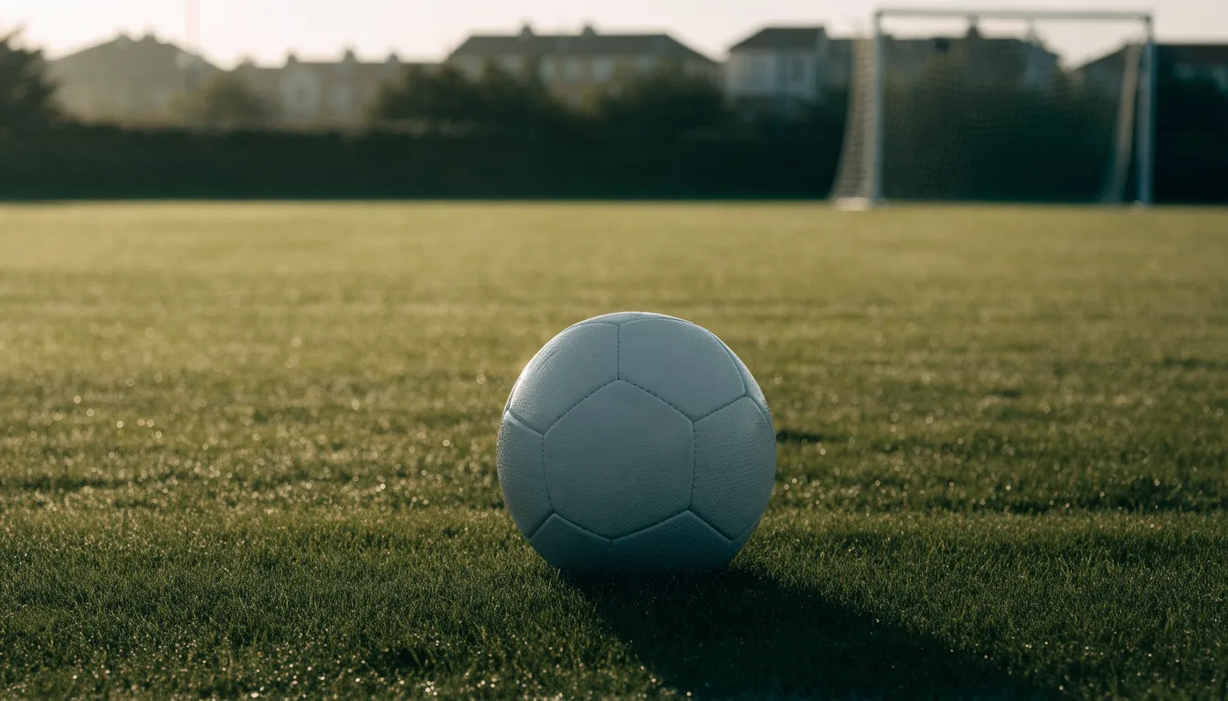 Ein Fußball auf dem Rasen eines leeren Sportplatzes am Rand einer Kleinstadt im Morgenlicht.
