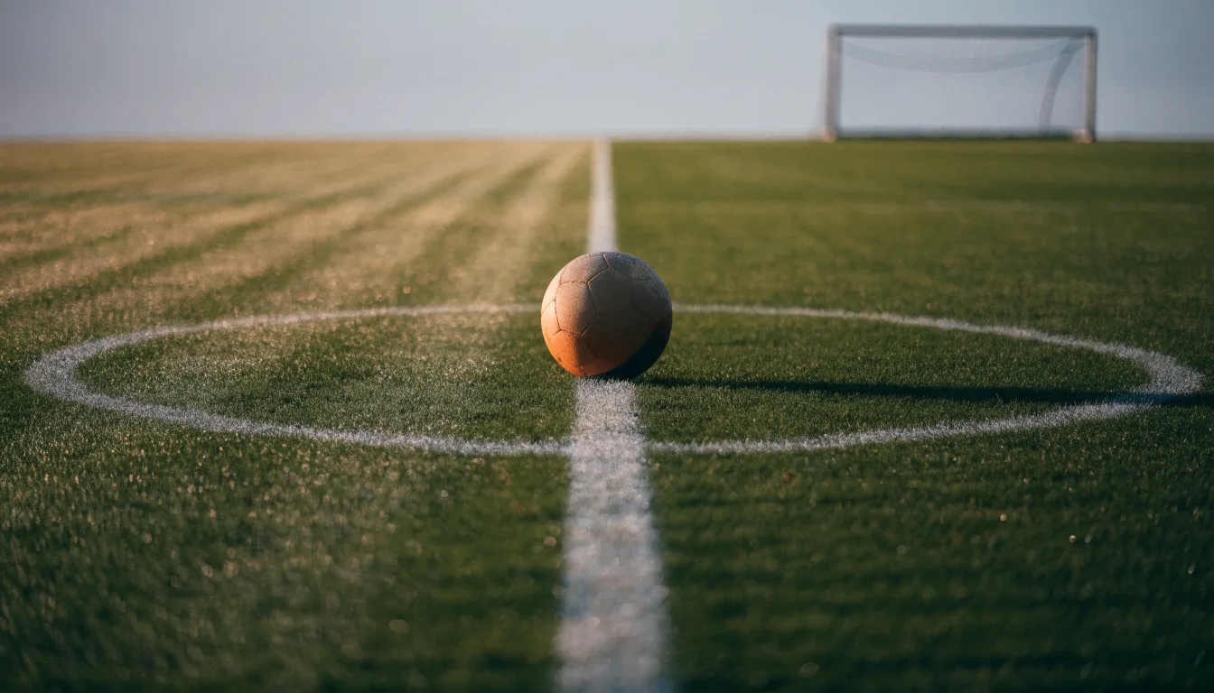 Ein Fußball ruht auf dem Mittelkreis eines leeren Rasenplatzes in der Morgendämmerung, Tau liegt auf dem Gras.