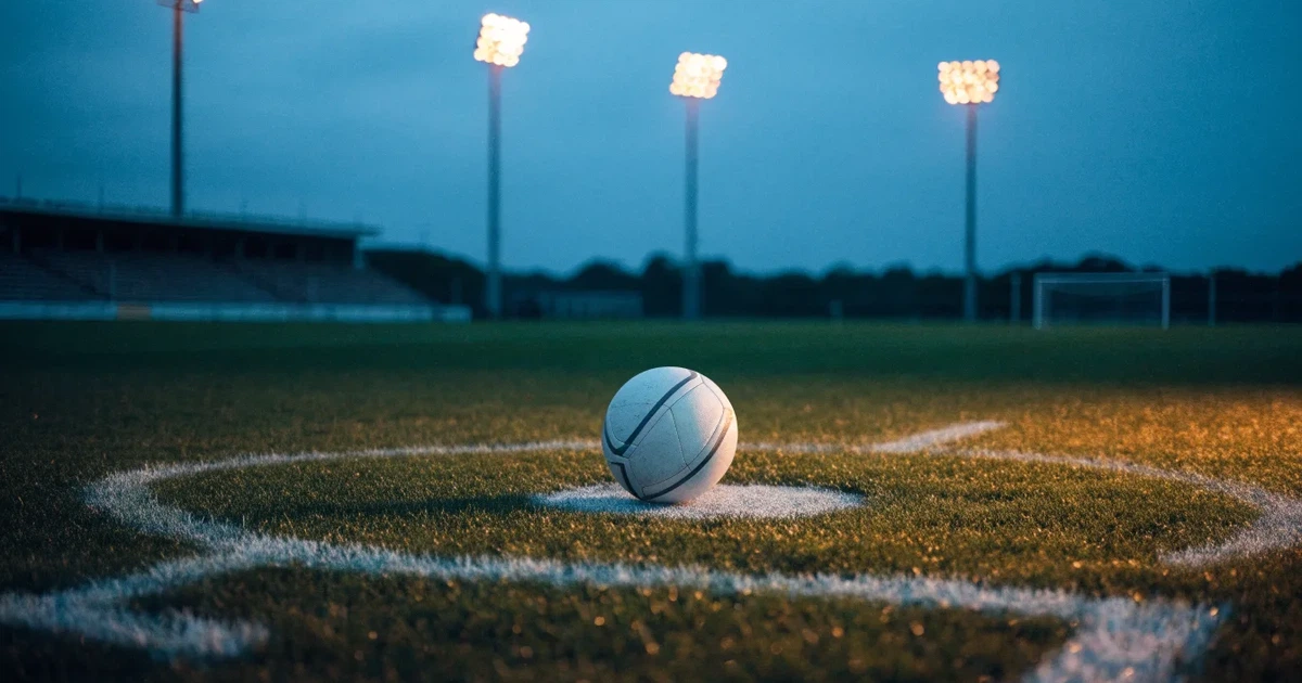 Ein Fußball liegt auf dem Anstoßpunkt eines leeren Rasenplatzes, im Hintergrund eine Tribüne und Flutlichtmasten in der Abenddämmerung.