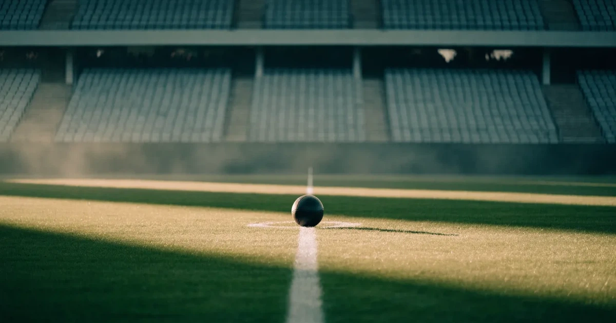 Ein Fußball liegt auf dem Anstoßpunkt eines Rasenplatzes, dahinter leere Tribünen im Licht des frühen Morgens.