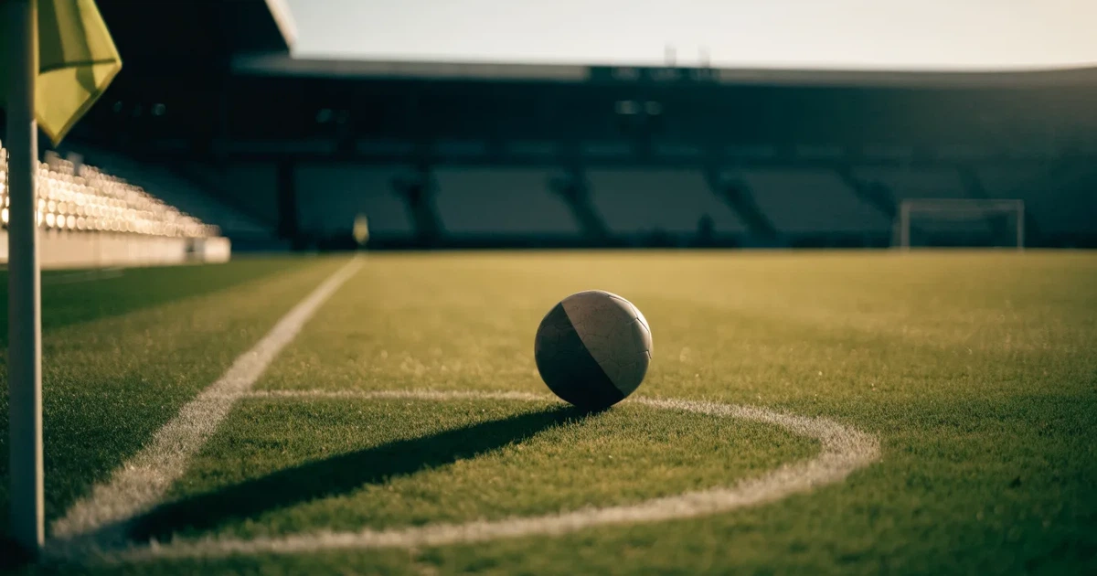 Ein Fußball liegt am Viertelkreis an der Eckfahne eines Rasenplatzes, im Hintergrund leere Tribünen in warmem Abendlicht.