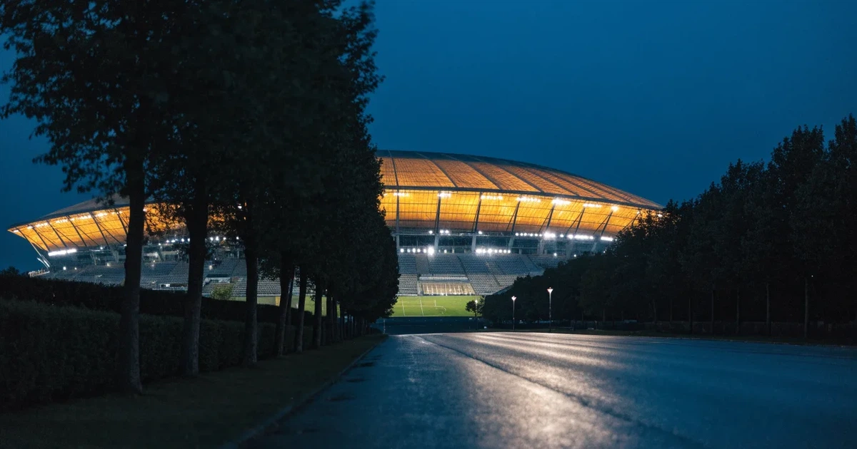 Ein Fußballstadion bei Nacht aus großer Entfernung, hell erleuchtet unter Flutlicht, davor eine dunkle Straße und Bäume.