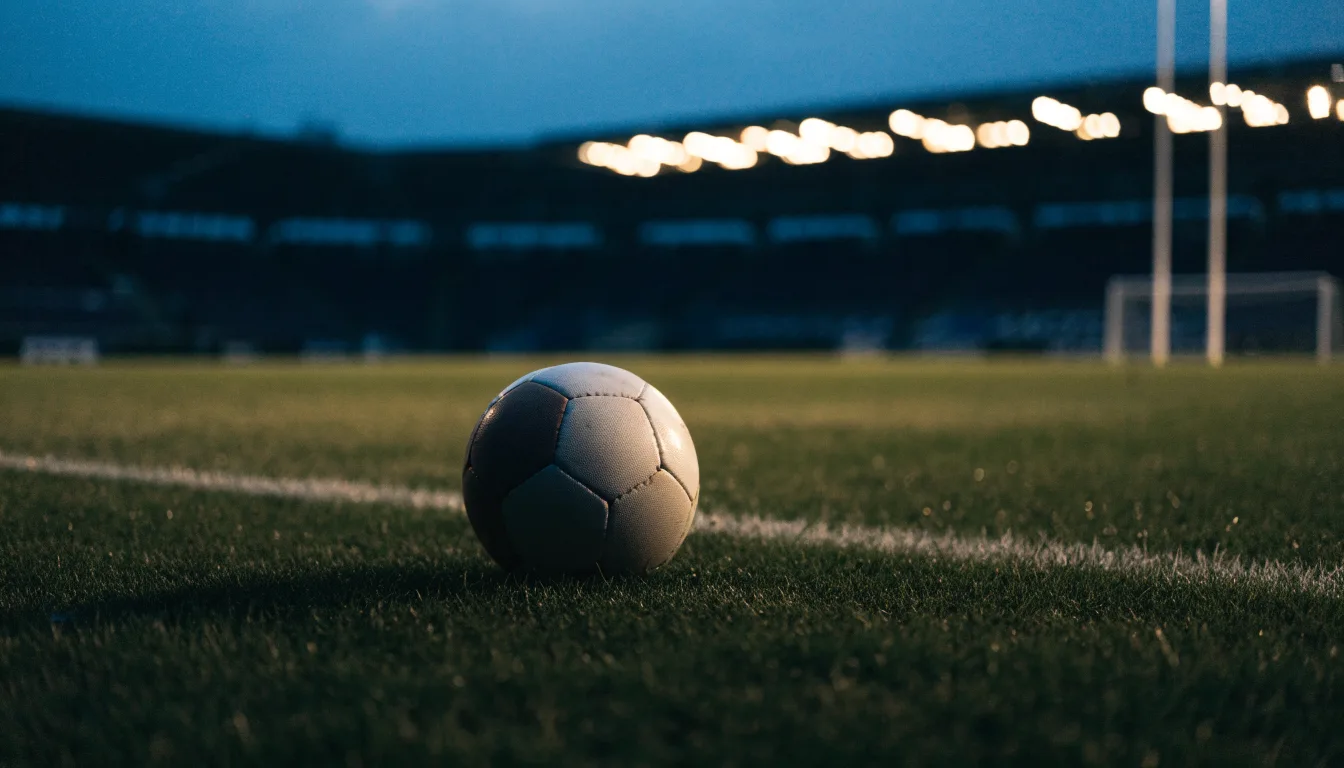 Ein Fußball liegt auf einem Rasenplatz in der Dämmerung, im Hintergrund ein ferner Stadionrand unter Flutlicht.